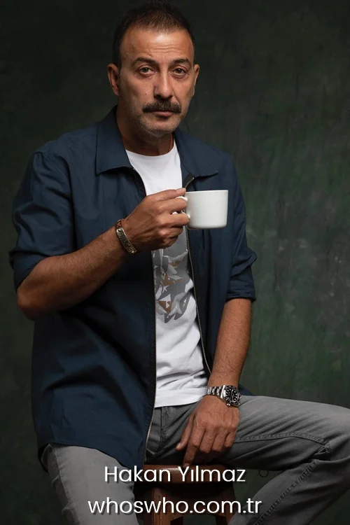 Professional studio portrait of Turkish actor Hakan Yılmaz sitting on a wooden stool and holding a white coffee cup, wearing an open dark blue zip-up shirt over a graphic white t-shirt, grey jeans, a silver luxury watch, and a metallic bracelet against a textured dark green background.