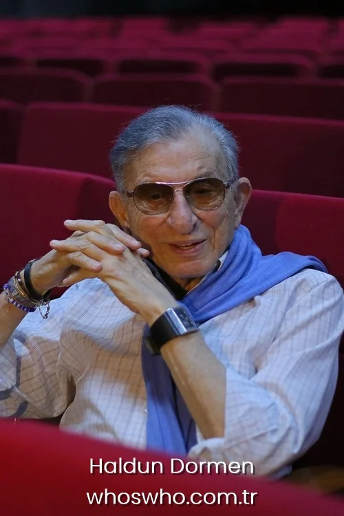 Legendary Turkish theater director Haldun Dormen smiling with sunglasses and a blue scarf in red theater seats.