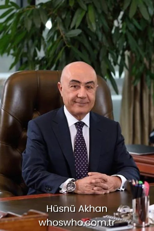 Turkish businessman Hüsnü Akhan smiling and clasping hands at his office desk in a dark suit.