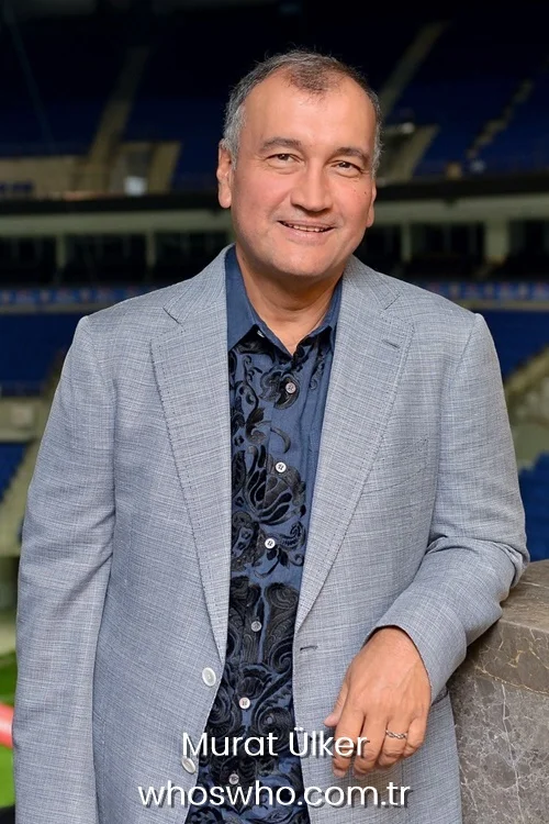 Turkish businessman Murat Ülker smiling in a grey blazer and patterned blue shirt at a sports arena.