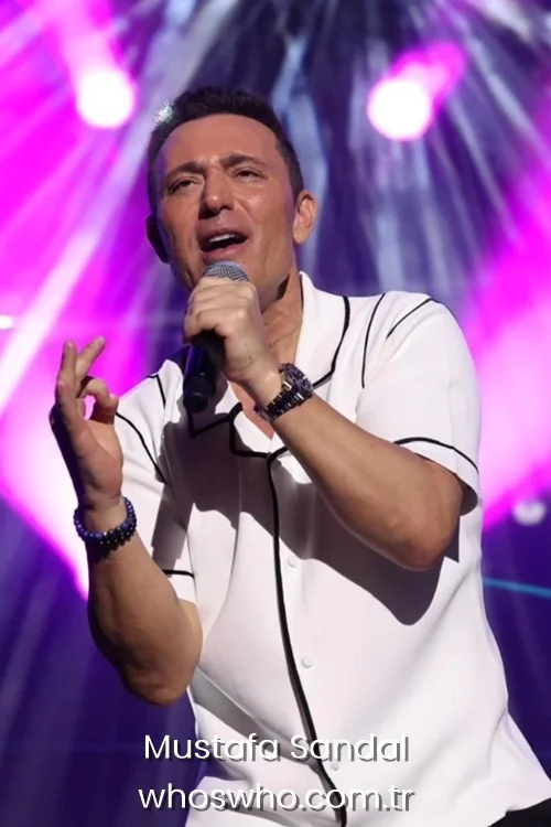 Turkish pop artist Mustafa Sandal singing into a microphone on stage under purple concert lights.