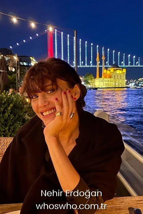 Turkish actress Nehir Erdogan smiling at an outdoor restaurant next to the Bosphorus sea with the illuminated Ortakoy Mosque and bridge in the background at night