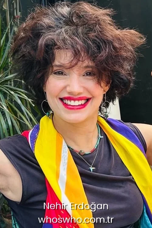 Turkish actress Nehir Erdogan with short messy brown hair smiling brightly while wearing a colorful yellow scarf and necklaces