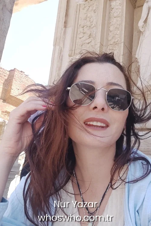 Close-up travel selfie of Turkish actress Nur Yazar smiling warmly outdoors, featuring her wind-blown brown hair, dark hexagonal sunglasses, layered necklaces, and a light blue jacket against a background of ancient stone ruins.