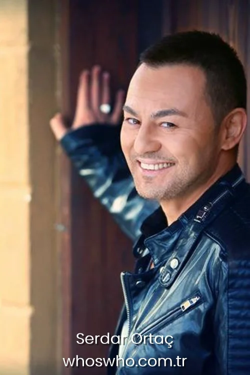 Turkish pop star Serdar Ortaç smiling in a black leather jacket while leaning against a wooden door.