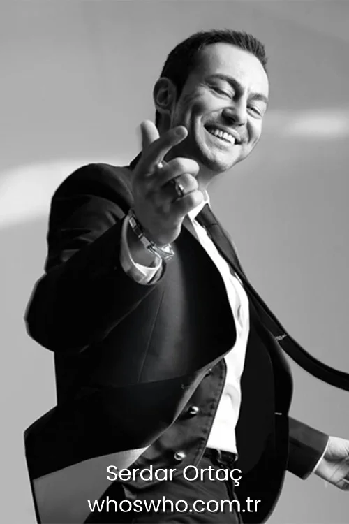 Black and white portrait of a young Turkish pop singer Serdar Ortaç smiling and gesturing happily in a suit.