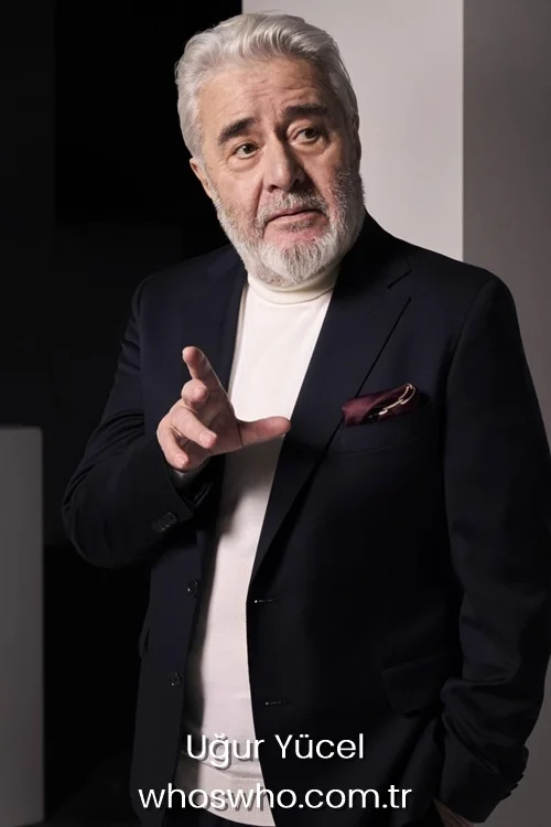 Turkish actor Uğur Yücel gesturing with his hand and looking to the side while wearing a dark navy blue blazer, a white turtleneck sweater, and a burgundy pocket square