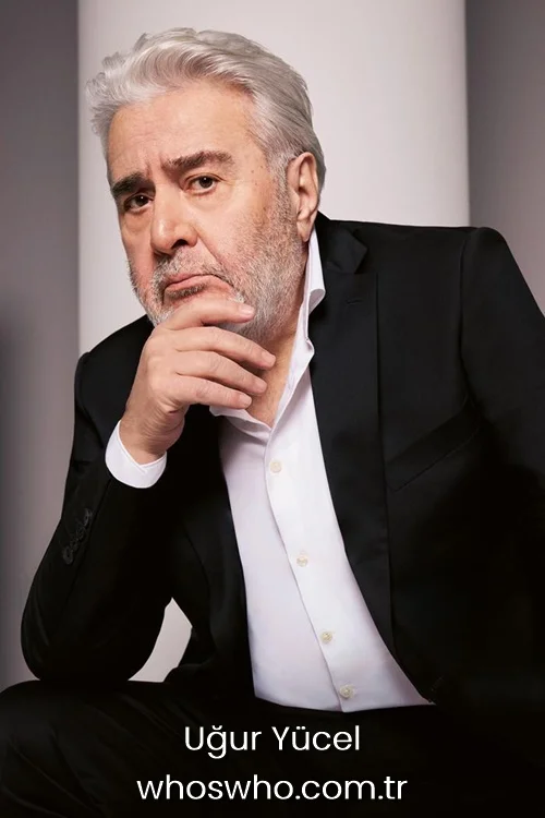 Turkish actor and director Uğur Yücel posing in a sharp black suit and open-collar white shirt, resting his chin on his hand with a pensive look.