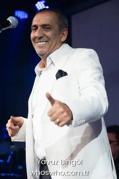 Yavuz Bingol smiling in a white suit holding a thumbs up on stage.
