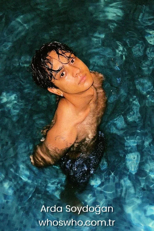 High-angle portrait of Crush boy band member Arda Soydoğan swimming in a pool, looking up at the camera with wet dark hair, showcasing a subtle arm tattoo through the clear blue water.