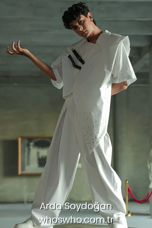 Full body portrait of Crush boy band member Arda Soydoğan posing dynamically indoors, showcasing his dark curly hair and wearing an avant-garde short-sleeved white shirt with black leather buckles and small rhinestone embellishments paired with high-waisted pleated wide-leg trousers and white shoes.