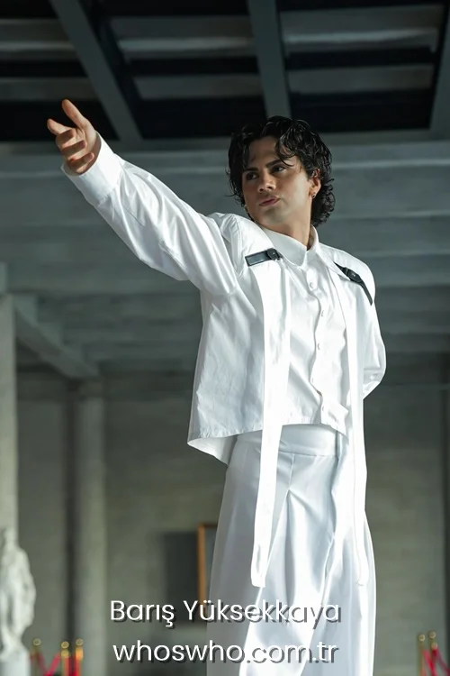 Medium shot of Crush boy band member Barış Yüksekkaya with wet-look curly dark hair posing dynamically with one arm extended, wearing an avant-garde white outfit featuring a long-sleeved cropped shirt with black leather strap details and matching high-waisted wide-leg trousers.
