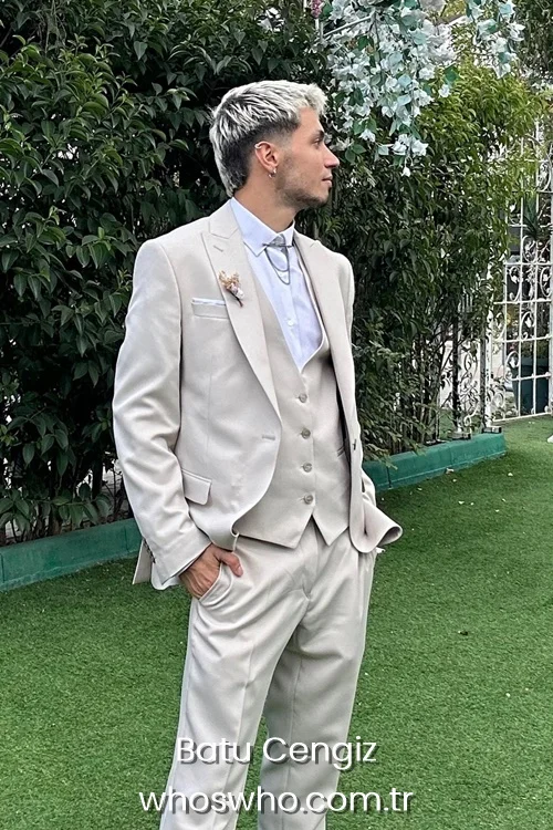 Side profile shot of Crush boy band member Batu Cengiz posing outdoors on a green lawn, showcasing his two-tone bleached blonde and dark hair, wearing an elegant cream-colored three-piece suit with a matching vest, a white dress shirt, and a silver collar chain.