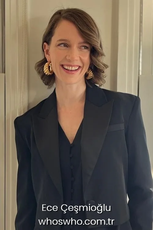 Actress Ece Cesmioglu smiling in a black blazer jacket and gold hoop earrings.