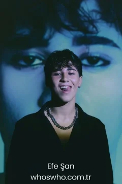 Efe Şan family Official close-up promotional portrait of Crush boy band member Efe Şan smiling playfully and sticking his tongue out, wearing an open black shirt and a heavy silver chain necklace under dramatic lighting with a large artistic projection of eyes in the blue background.