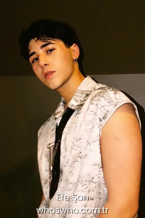 Close-up portrait of Crush boy band member Efe Şan looking at the camera, wearing a sleeveless white button-up shirt with a grey marble pattern, a dark tie styled with a silver safety pin and chains, and a small silver hoop earring.