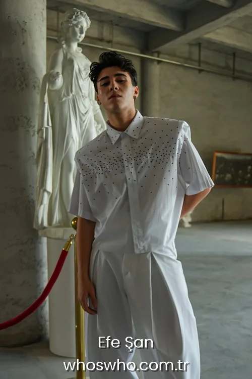 Medium shot of Crush boy band member Efe Şan posing confidently indoors next to a white marble classical statue, wearing an avant-garde short-sleeved white shirt embellished with small black studs across the chest, paired with matching white high-waisted pleated trousers.