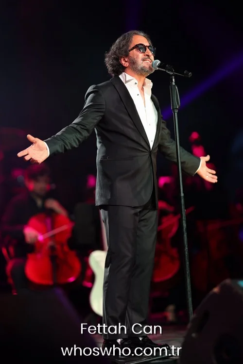 Turkish singer Fettah Can performing live on stage with a microphone in a black tuxedo suit during a concert with an orchestra background