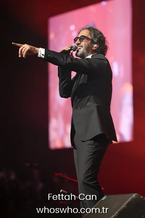 Turkish musician Fettah Can singing with microphone and pointing finger on stage with large red LED screen background