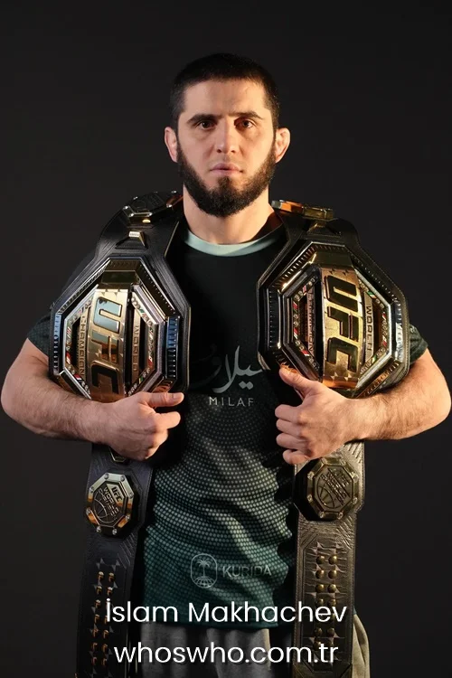 MMA fighter Islam Makhachev posing with two gold UFC championship belts over his shoulders.