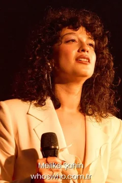 Turkish pop singer Melike Şahin singing with microphone on stage in a white blazer jacket with curly hair hair under warm concert lighting