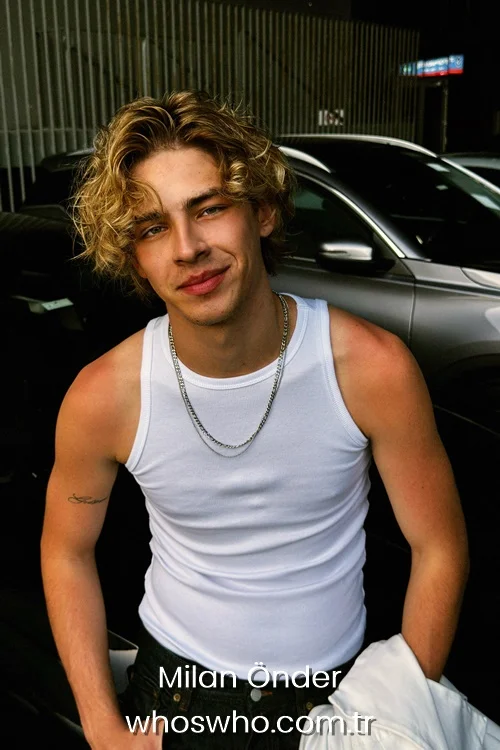 A candid medium portrait of Crush boy band member Milan Önder smiling warmly at the camera outdoors, showcasing his wavy blonde hair, wearing a white ribbed tank top, black jeans, a thin silver chain necklace, and holding a white jacket while standing in front of cars.