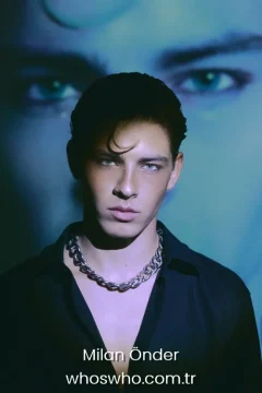 Milan Önder age Official close-up promotional portrait of Crush boy band member Milan Önder posing in an open black shirt and a thick silver chain necklace, featuring styled dark hair with a single curl on his forehead, looking directly at the camera under dramatic lighting with a large artistic projection of eyes in the blue background.