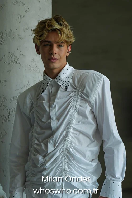 Medium shot of Crush boy band member Milan Önder posing indoors against a textured wall, showcasing his blonde wavy hair, wearing a unique avant-garde white long-sleeved shirt with gathered ruching details on the front and black polka dots on the collar and cuffs.