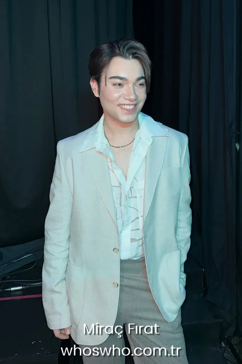 Medium shot of Crush boy band member Miraç Fırat smiling warmly backstage, featuring his side-parted dark hair, wearing a light beige blazer over a white patterned button-up dress shirt, a delicate gold chain necklace, and brown trousers against a dark curtain background.