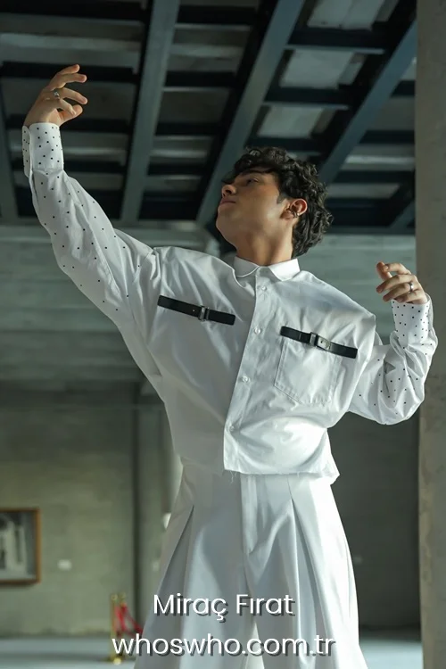 Medium shot of Crush boy band member Miraç Fırat posing gracefully with one arm raised and head tilted back, wearing an avant-garde white outfit featuring a long-sleeved shirt with black leather strap buckle details on the chest, patterned sleeves, and high-waisted pleated trousers.