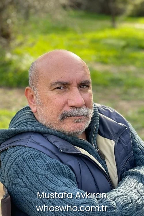 Close-up outdoor portrait of veteran Turkish actor Mustafa Avkıran looking at the camera with a calm expression, showcasing his thick grey mustache and beard, wearing a heavy teal cable-knit hooded sweater under a dark blue puffer vest against a blurred green nature background.