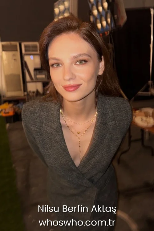 Actress Nilsu Berfin Aktas smiling in a gray blazer outfit with gold necklaces.