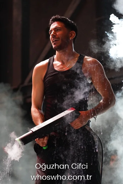 Dynamic medium shot of Crush boy band member Oğuzhan Çifçi performing passionately on stage, wearing a fitted black ribbed tank top and a thin gold chain necklace, holding a smoke cannon blaster that emits white smoke effects around him.