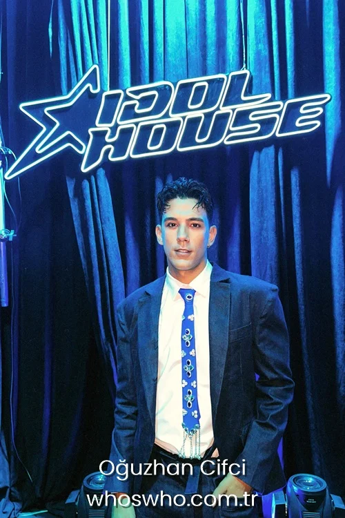 Medium shot of Crush boy band member Oğuzhan Çifçi posing at the Idol House event, wearing a dark blue suit jacket over a white dress shirt, accessorized with a unique bright blue necktie decorated with jewels and fringe, standing in front of a blue velvet curtain with a neon Idol House sign.