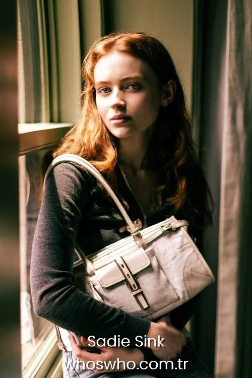 Actress Sadie Sink posing with a white shoulder handbag by a window for a lifestyle feature.