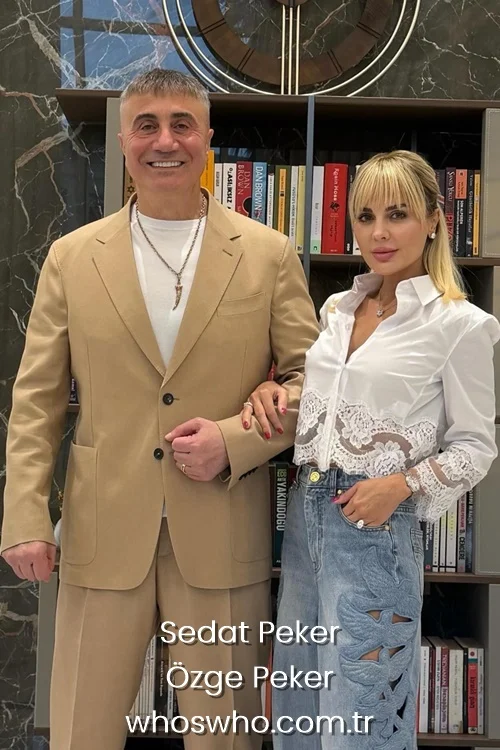 A professional indoor portrait of Sedat Peker wearing a formal beige suit jacket over a white t-shirt and his wife Özge Peker in a stylish white lace blouse and light blue embroidered denim pants, posing arm in arm next to a modern wooden library bookshelf.