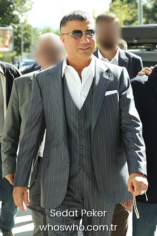 Medium shot of Sedat Peker walking outdoors surrounded by men, wearing a tailored dark grey pinstripe three-piece suit with a matching vest, an open white dress shirt, and rimless sunglasses while holding prayer beads in his hand.
