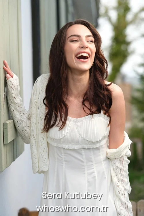 Candid medium shot of Turkish actress Sera Kutlubey laughing joyfully outdoors, showcasing her long wavy brown hair, wearing an elegant pleated white dress and a white knit cardigan slipping off her shoulder while leaning against a rustic wooden shutter.