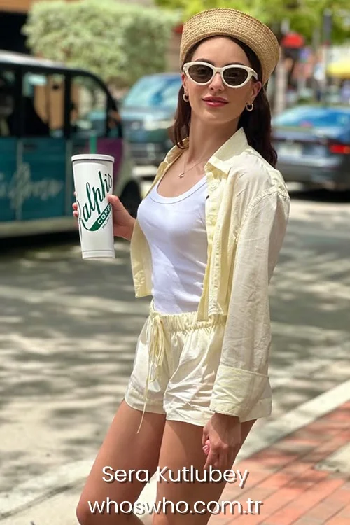 Full shot of Turkish actress Sera Kutlubey walking on a city street, holding a large white Ralph's Coffee cup, wearing a matching pastel yellow button-up shirt and shorts set over a white tank top, accessorized with a straw beret hat, white sunglasses, and gold hoop earrings.