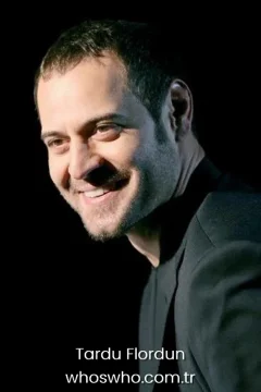 Side profile portrait of handsome Turkish actor Tardu Flordun smiling warmly, showcasing his short dark hair and slight stubble beard, wearing a formal black suit jacket against a solid black background.