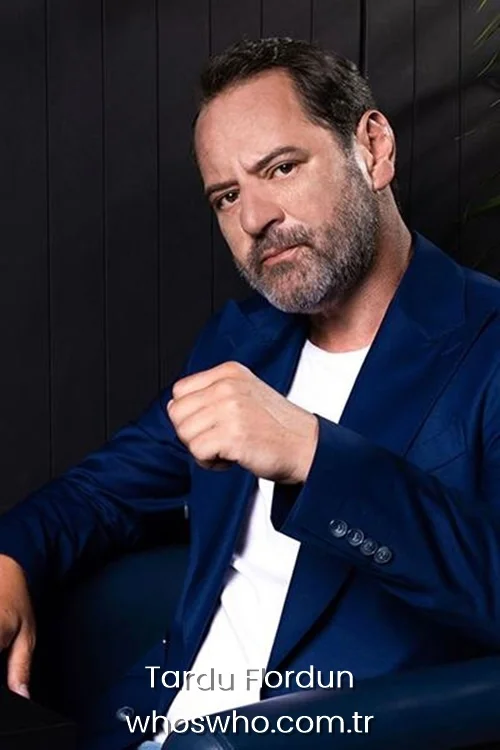 Medium shot of Turkish actor Tardu Flordun sitting in a chair with a serious expression and a raised fist, featuring his peppered grey beard and short dark hair, wearing a stylish royal blue blazer over a casual white t-shirt against a dark wood wall backdrop.