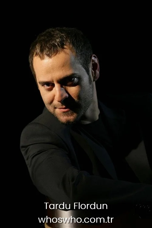 Close-up dramatic portrait of Turkish actor Tardu Flordun leaning forward and looking intently at the camera under artistic low-key lighting, featuring his short dark hair and a neat stubble beard, wearing a sharp black suit against a solid black background.