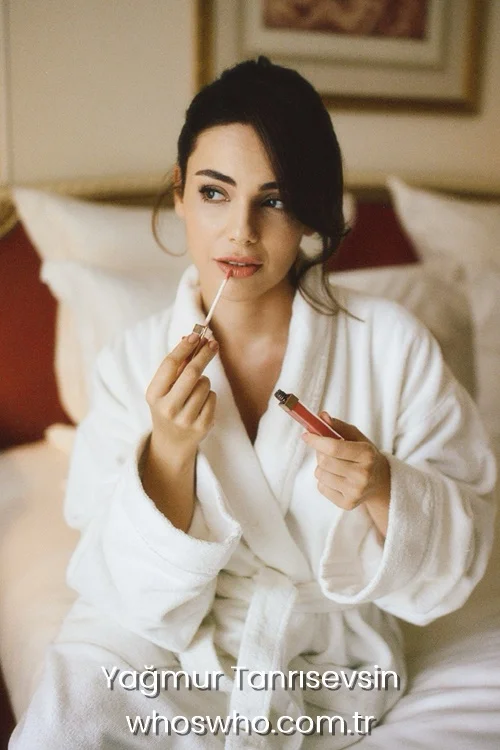 Turkish actress Yağmur Tanrısevsin sitting on a bed and applying lip gloss.